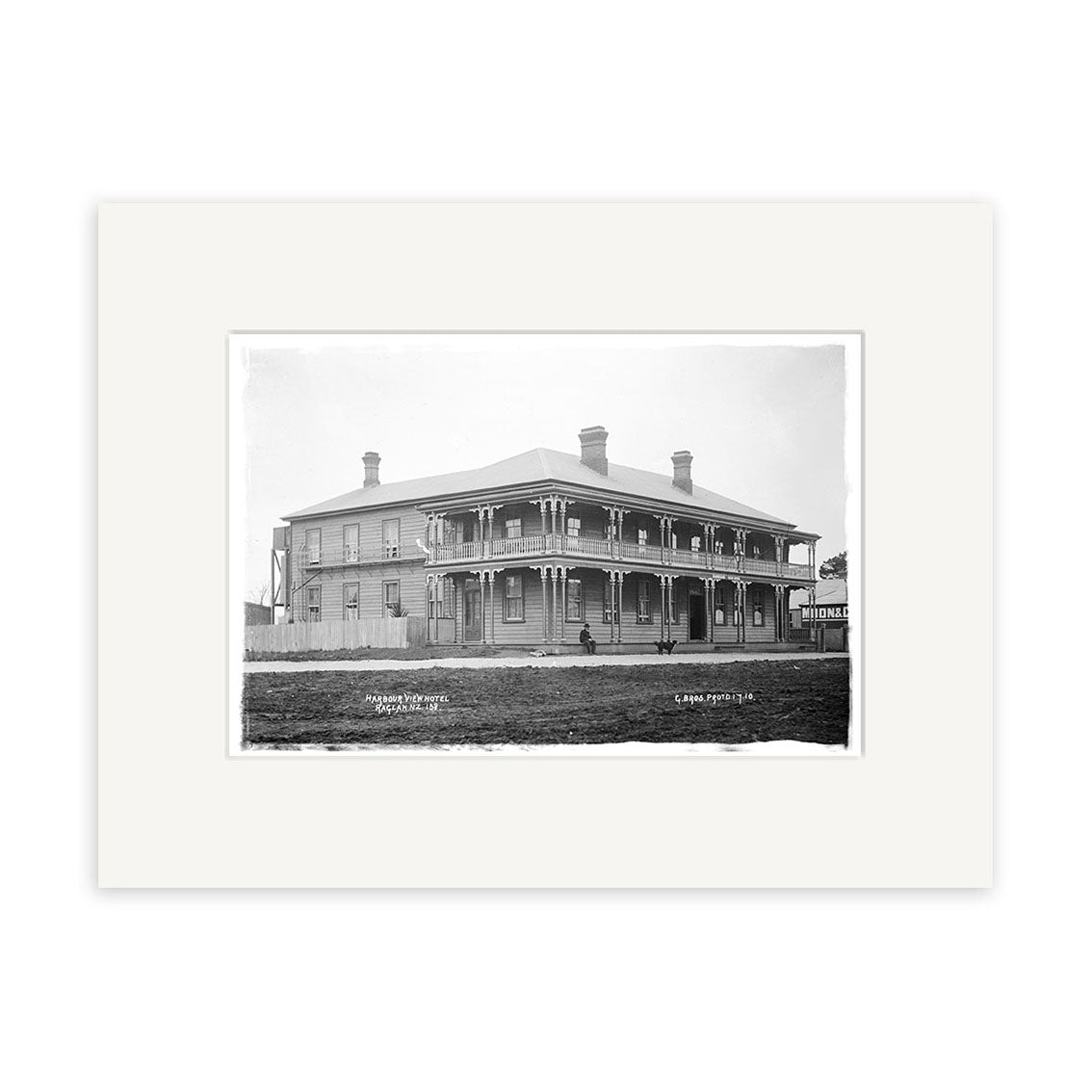Archival quality black and white photo print of Harbour View Hotel 1910 (HI179), showing a two-story wooden building with wraparound porch, chimneys, people on the steps, and a picket fence in New Zealand. Mounted with white mat.