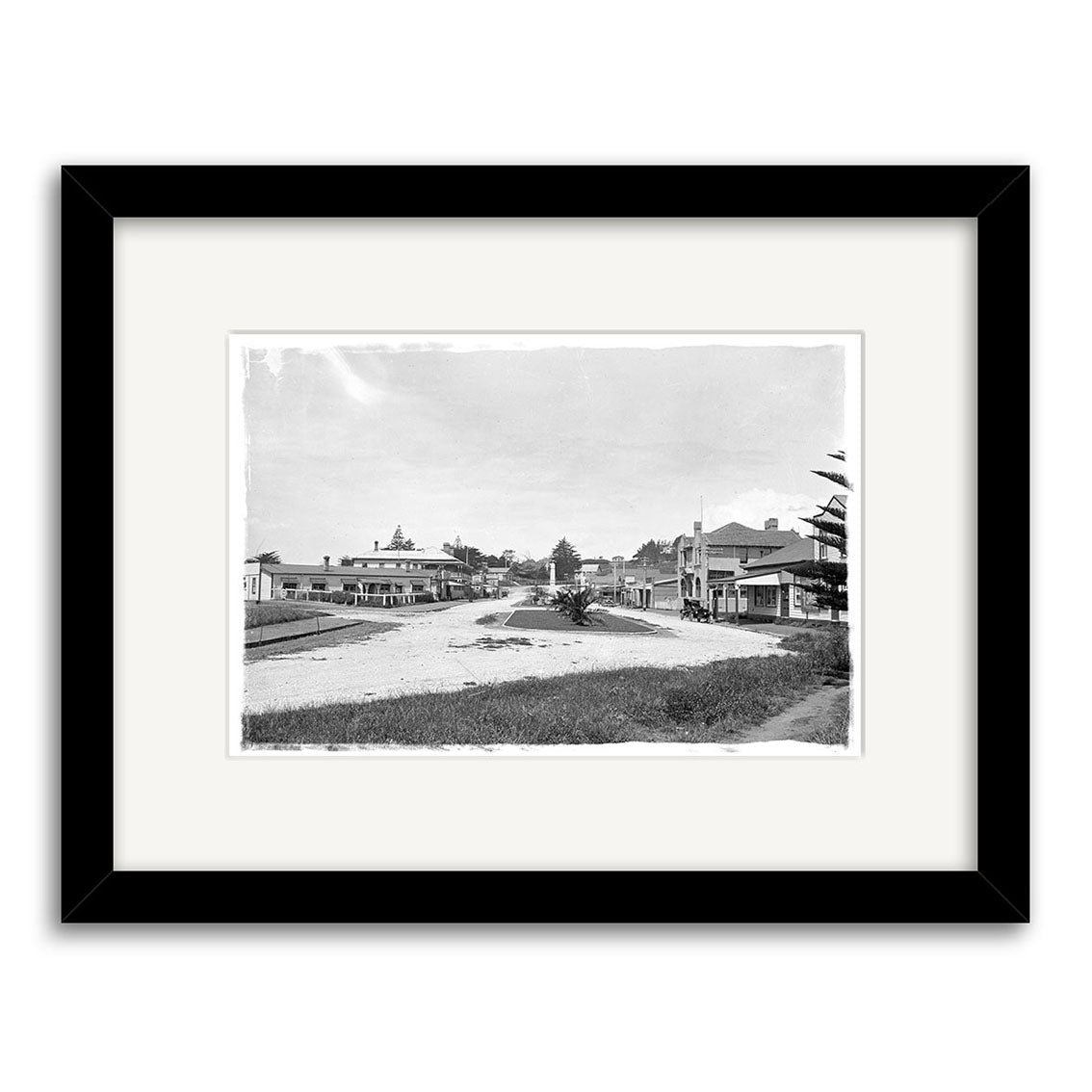 Archival quality black and white photograph, Main Street 1929 - HI180, in a black frame, depicting a quiet early 20th-century New Zealand street with low buildings, trees, and an unpaved road.
