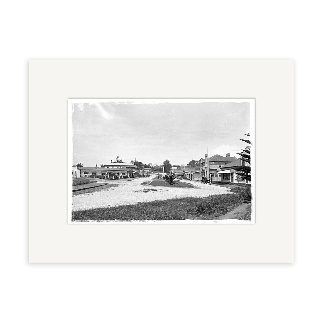 Main Street 1929 - HI180 is a black and white historical photographic print of a quiet, wide dirt street in a small New Zealand town with wooden buildings and trees. This archival-quality image features a white border on a white background.