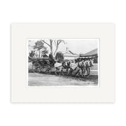 Black and white photo titled Raglan Mail Coach 1912 - HI181 shows a man on a horse-drawn carriage with four horses along a dirt road in New Zealand, with trees, houses, and a white picket fence visible in the background.