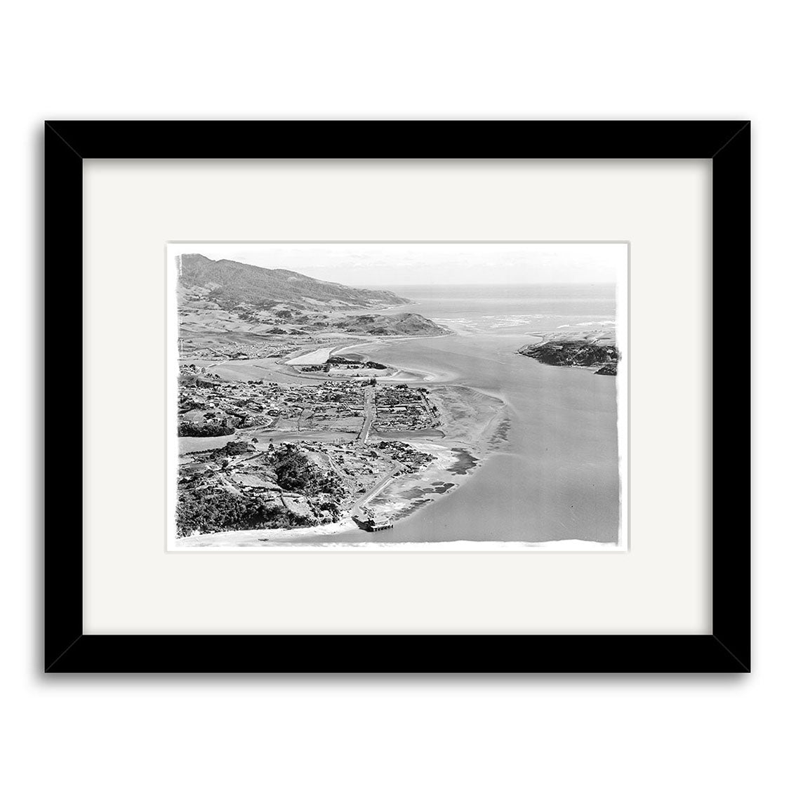 Black and white aerial photo Raglan 1957 - HI183 shows a New Zealand coastal landscape with river or bay, hills, and scattered buildings; framed with a white mat and black frame on archival matte paper.