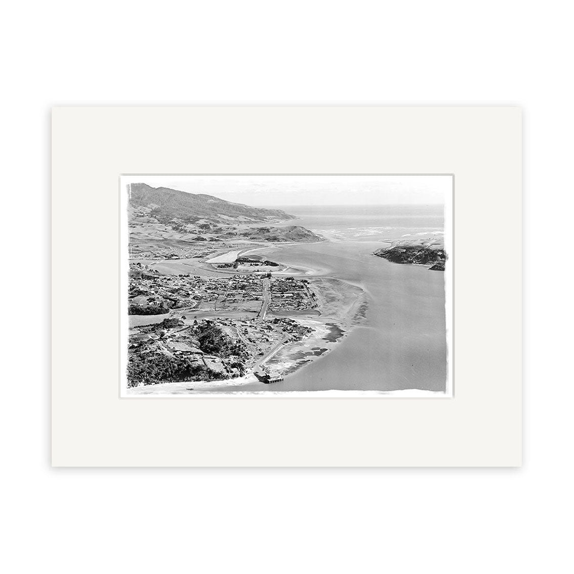 The Raglan 1957 - HI183 print displays a black and white aerial view of a New Zealand coastal landscape with mountains, water, and buildings, printed on archival quality matte paper for long-lasting preservation.