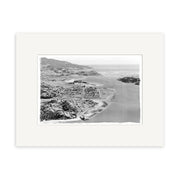 The Raglan 1957 - HI183 print displays a black and white aerial view of a New Zealand coastal landscape with mountains, water, and buildings, printed on archival quality matte paper for long-lasting preservation.