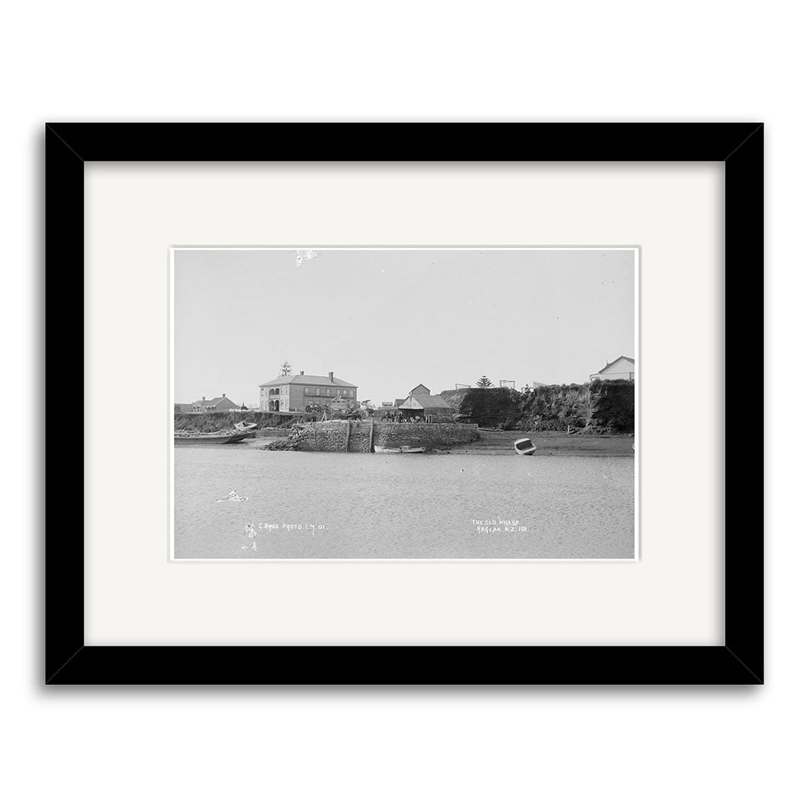 Archival-quality black-and-white photographic print, titled Raglan 1910 No.2 - HI185, features small boats by a New Zealand dock and calm water; presented in a black frame with white mat for display.