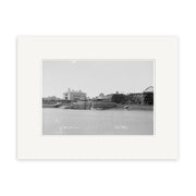 Black and white photo titled Raglan 1910 No.2 - HI185 shows a small harbor with boats by a dock and buildings in the background. The historic print has a white archival-quality mat border for preservation.