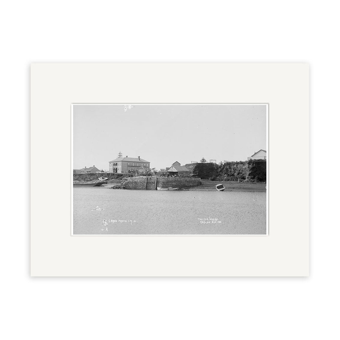 Black and white photo titled Raglan 1910 No.2 - HI185 shows a small harbor with boats by a dock and buildings in the background. The historic print has a white archival-quality mat border for preservation.
