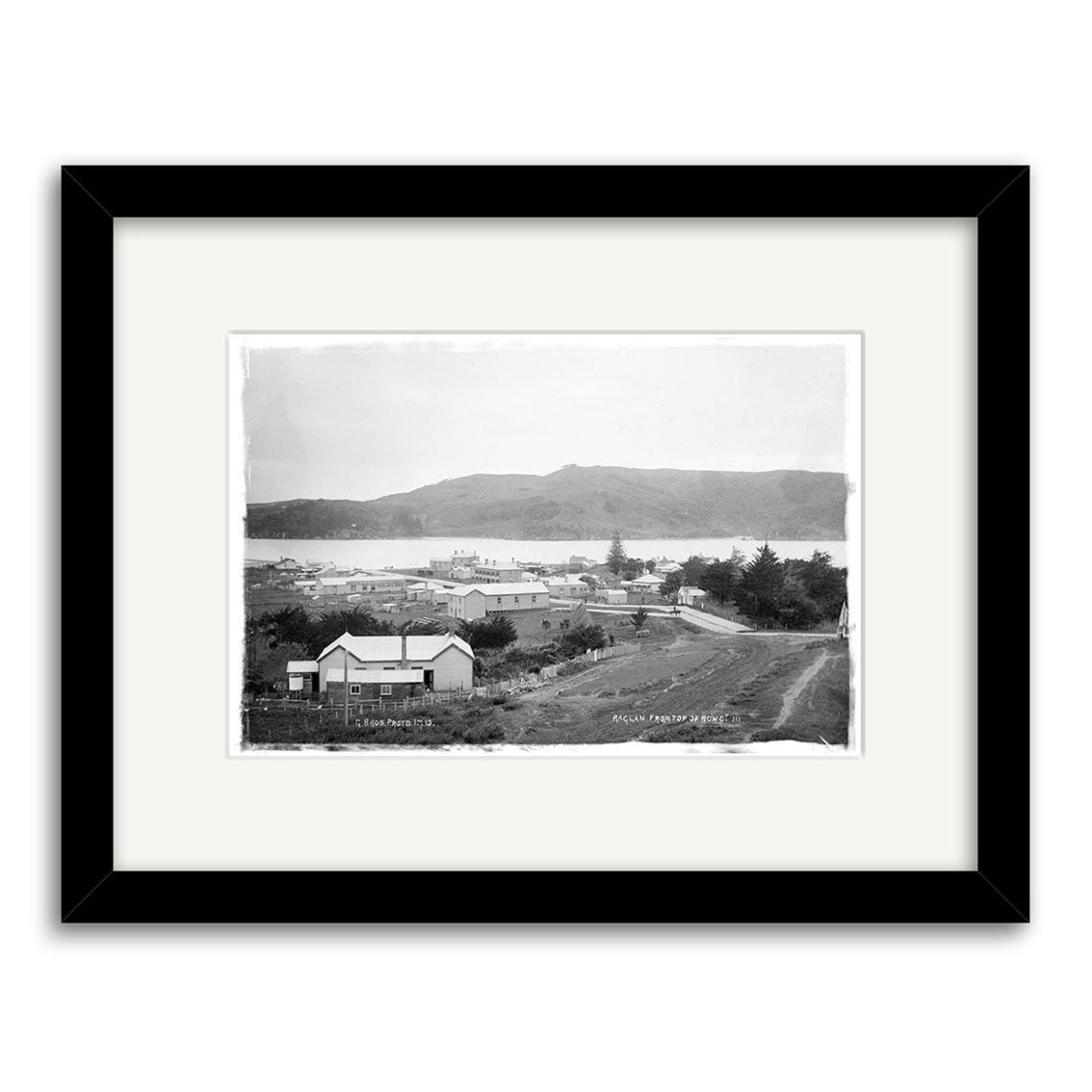 Archival quality black and white framed photograph, Raglan 1910 No.3 - HI186, featuring a small New Zealand town with scattered buildings, dirt roads, trees in the foreground, and hills with water in the background.