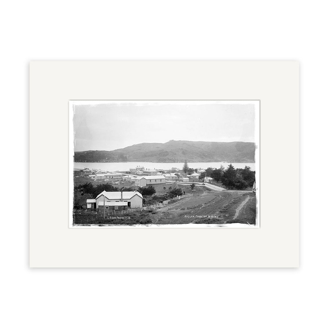 The Raglan 1910 No.3 - HI186 black and white historical photographic print depicts a small New Zealand town with buildings, a dirt road, trees, and hills. It is bordered by an archival quality white mat.