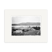 The Raglan 1910 No.3 - HI186 black and white historical photographic print depicts a small New Zealand town with buildings, a dirt road, trees, and hills. It is bordered by an archival quality white mat.