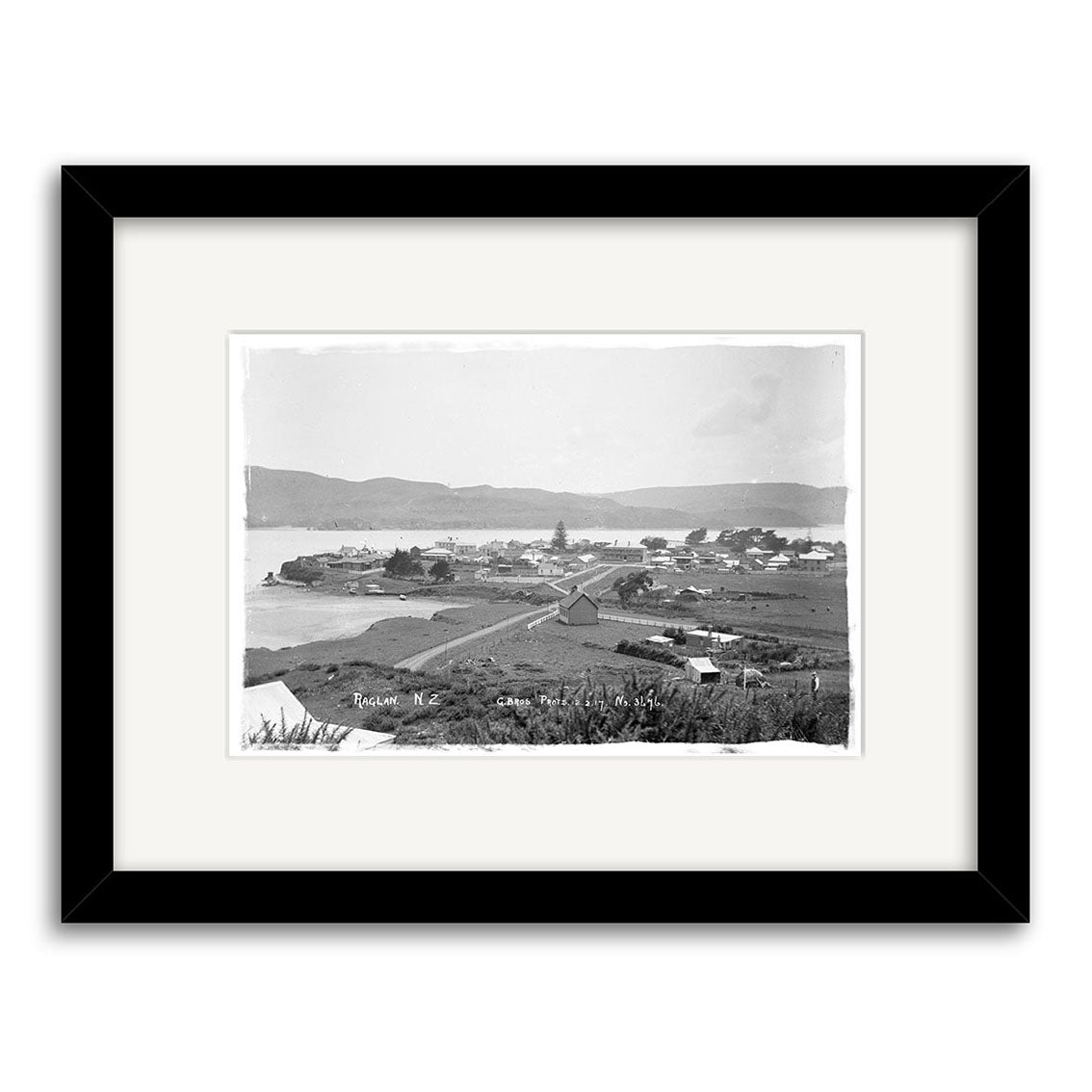 Black and white framed Historical Photographic Print titled Raglan 1917 - HI187, showing a coastal town with buildings, road, grassy fields, water, and hills. Printed on matte paper; “Russell N.Z.” appears in the lower left corner.