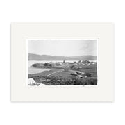 Archival quality black and white photographic print of Raglan 1917 (HI187), showing a coastal town with houses, open land, road, water, and hills in the background. Framed with a white mat.