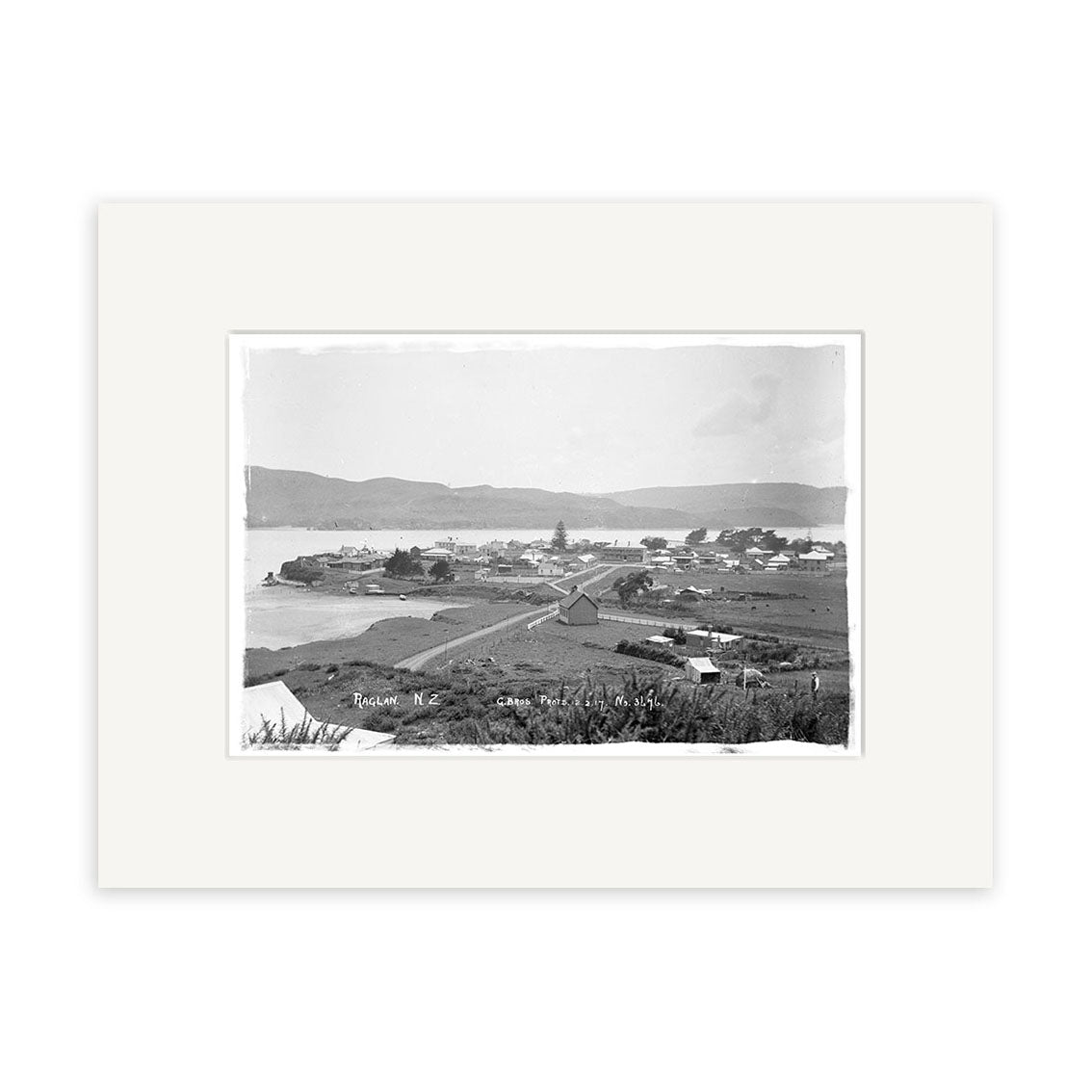 Archival quality black and white photographic print of Raglan 1917 (HI187), showing a coastal town with houses, open land, road, water, and hills in the background. Framed with a white mat.
