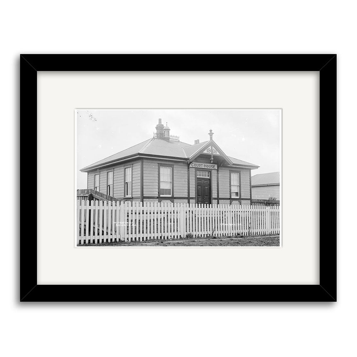 Black and white photo of the Raglan Court House 1910 (HI188), a small wooden building with “Court House” sign, picket fence, in a black frame with white mat. Historical photographic print made in New Zealand.