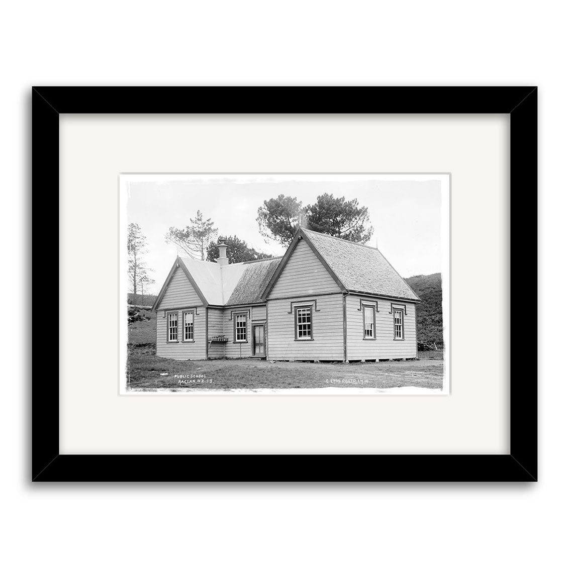 Archival-quality black and white framed print of Raglan Public School 1910 (HI189), featuring a single-story wooden building with a gabled roof, multiple windows, and trees behind; displayed with a white mat and black frame. Made in New Zealand.
