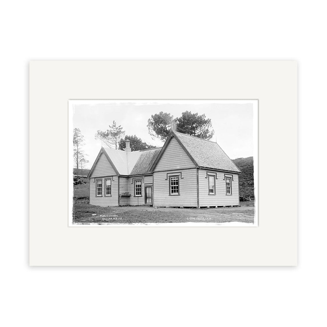 A black and white historical photographic print titled Raglan Public School 1910 - HI189 shows a single-story wooden house with a gabled roof set amid grassy New Zealand surroundings, mounted in an archival quality white mat.