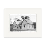 A black and white historical photographic print titled Raglan Public School 1910 - HI189 shows a single-story wooden house with a gabled roof set amid grassy New Zealand surroundings, mounted in an archival quality white mat.