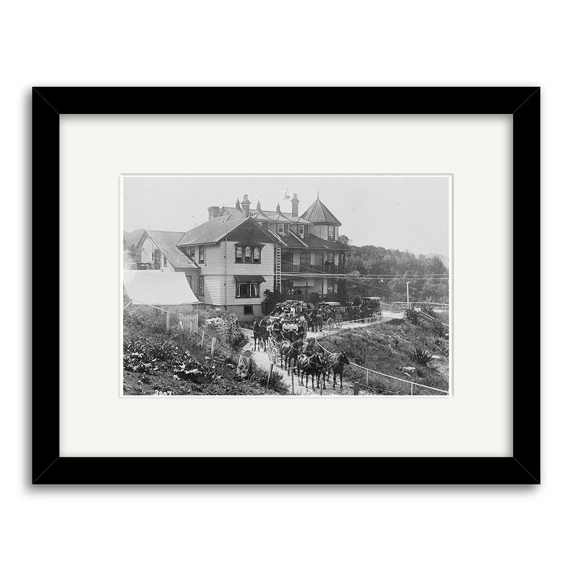 Black-and-white photographic print titled Hotel Waitomo 1910 - HI190, showing a large Victorian-style house with a wraparound porch and three horse-drawn carriages, framed with a white mat and black frame. Archival quality, evoking historic New Zealand charm.