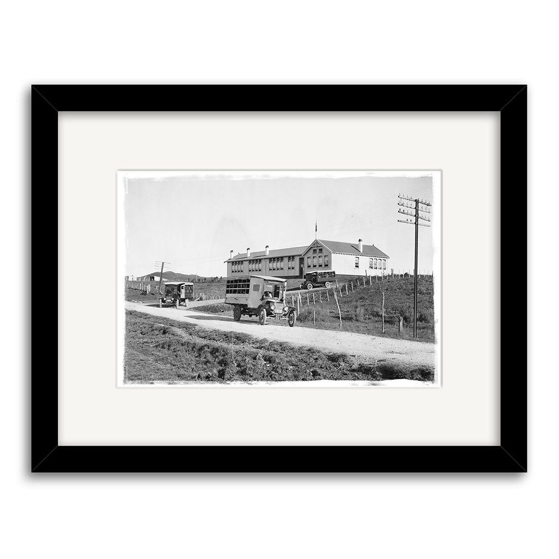 Archival black-and-white print titled Piopio School Waitomo 1924 No.2 - HI192 shows early 20th-century delivery trucks on a dirt road by a fenced building with an American flag; white mat and black frame included.