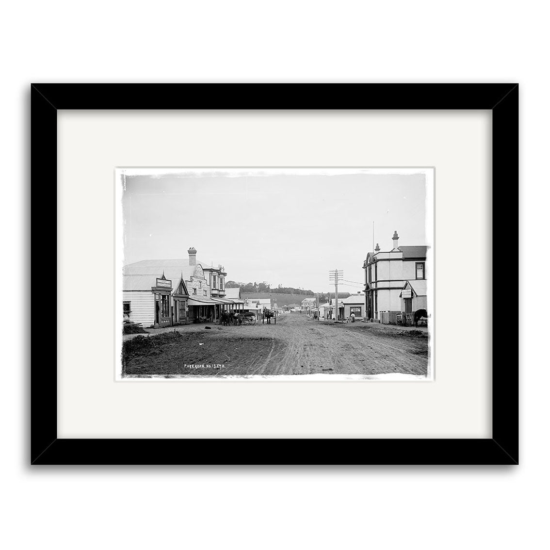 King Street 1909 - HI194: Black and white historical photo of a wide dirt road with wooden buildings, utility poles, and townspeople. Framed in black with white mat on archival-quality matte paper.