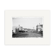 King Street 1909 - HI194: Black and white historical photographic print on matte archival paper shows a dirt road with wooden buildings and utility poles, capturing the spirit of a small town in the early 20th century.