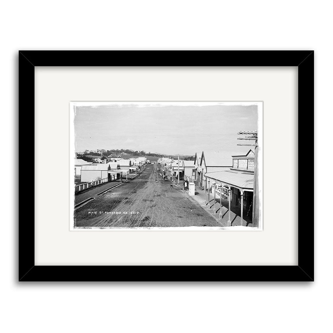 Archival-quality framed print: Main St Pukekohe 1900s - HI195 shows a wide dirt street lined with wooden buildings and shops in New Zealand, viewed from above. Few people appear on the mostly empty street. White mat, black frame.