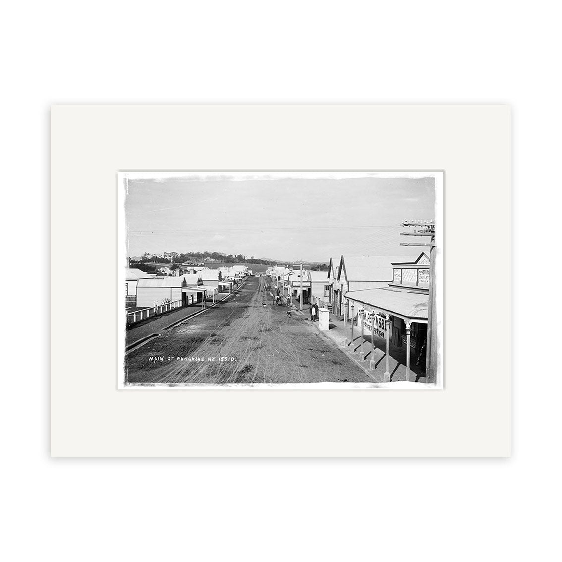 Archival black and white photographic print, Main St Pukekohe 1900s - HI195, shows a wide dirt street lined with small buildings and shops in New Zealand, with a few people strolling the sidewalks.