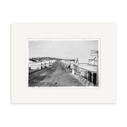 Archival black and white photographic print, Main St Pukekohe 1900s - HI195, shows a wide dirt street lined with small buildings and shops in New Zealand, with a few people strolling the sidewalks.