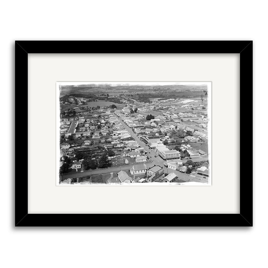 Pukekohe 1946 - HI196: A black and white aerial photograph of a town with small buildings, roads, and trees. Printed on archival quality matte paper and framed with a wide white mat in a simple black frame.