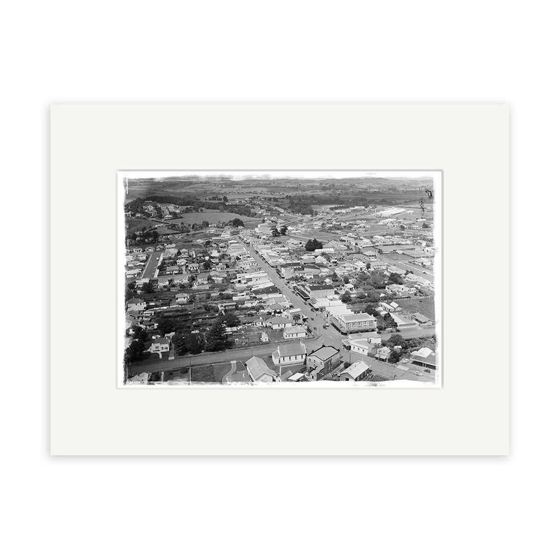 Black and white aerial photograph titled Pukekohe 1946 - HI196 shows a small town with houses, buildings, and roads amid open countryside. This historical print is mounted with a wide white mat.