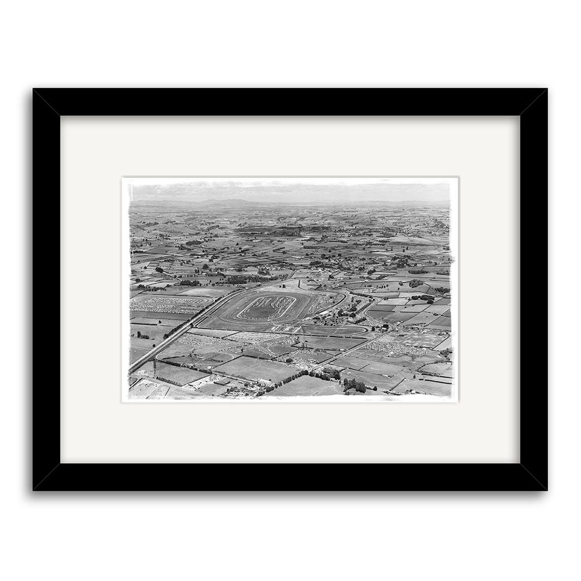 Pukekohe 1963 - HI197: Black and white aerial photo of a large rectangular racetrack amid open fields and scattered buildings, presented as a historical photographic print in a black frame with white mat on archival matte paper.