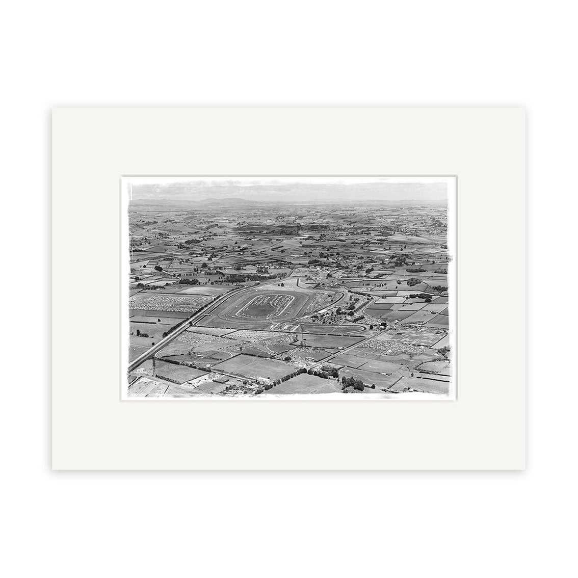 The Pukekohe 1963 - HI197 is a black and white aerial photo print of a racetrack amid fields and farmland, with scattered buildings and roads. The historical image comes matted with a plain white border.