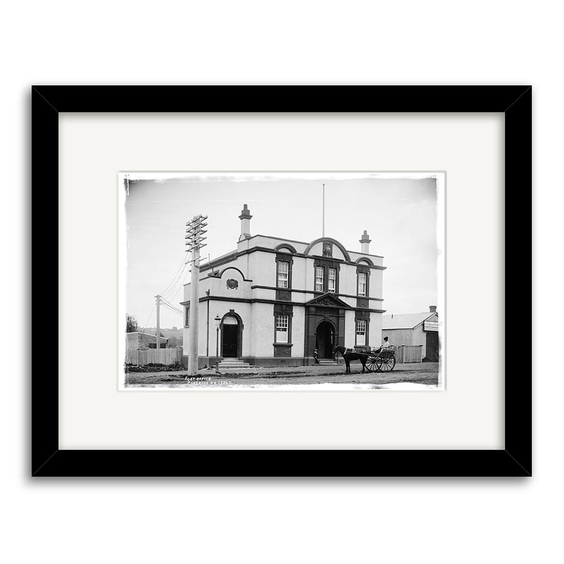 Archival quality print of the Pukekohe Post Office 1909 (HI198) in New Zealand, showing a two-story building with arched windows, central entrance, black frame, horse-drawn carriage, and utility poles in front.