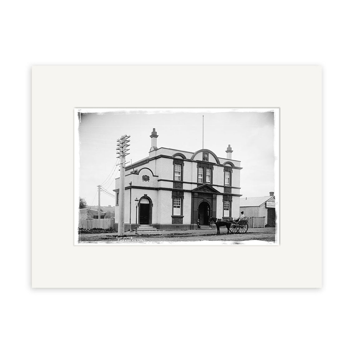 Black-and-white archival print: Pukekohe Post Office 1909 (HI198). Features a two-story building with arched windows, horse-drawn carriage, and telephone poles. Ideal for collectors of New Zealand history.