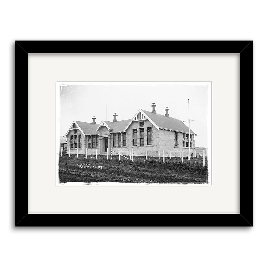 A black and white photographic print titled Pukekohe Primary 1910 - HI199 features a single-story, gabled-roof building with multiple chimneys, set within a fenced grassy area and framed with a black border and white mat.