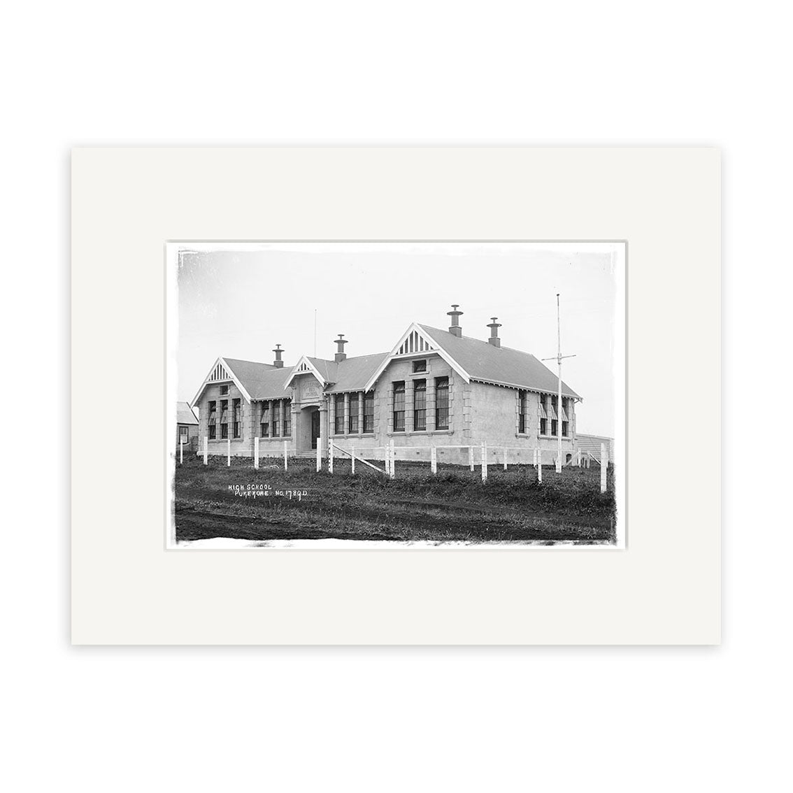 Pukekohe Primary 1910 - HI199: Black-and-white photo of a single-story building with large windows, gabled roofs, and chimneys, surrounded by a white fence and grass, mounted on archival quality matte paper.