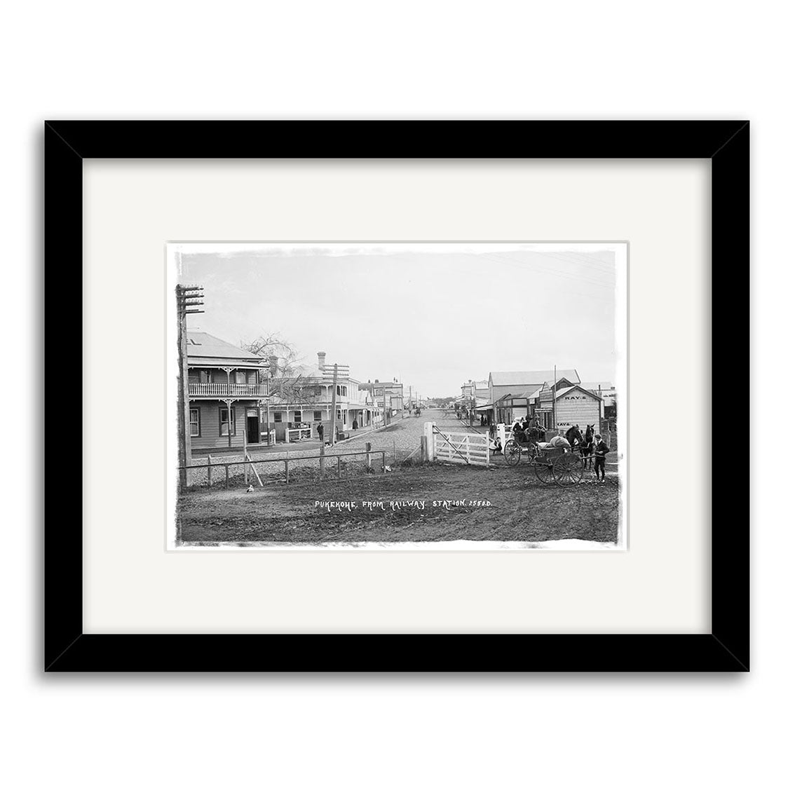 Archival quality historical photographic print of Pukekohe 1910 - HI200, depicting a black-and-white street scene with early 20th-century buildings, utility poles, people, a horse-drawn carriage, and a gate in the foreground.