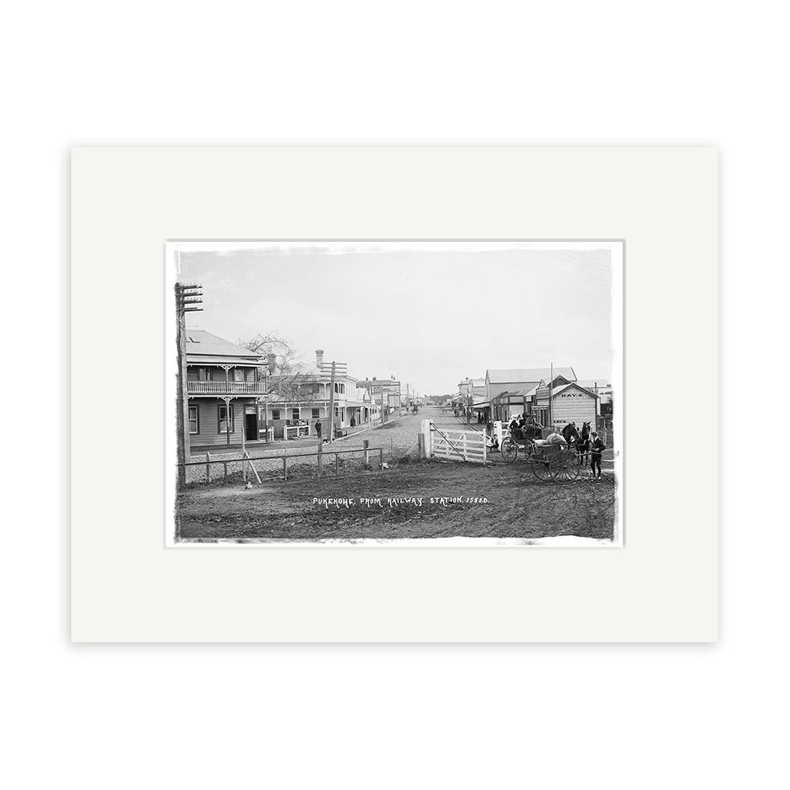 Archival quality photographic print titled Pukekohe 1910 - HI200 featuring a small New Zealand town street with wooden buildings, horse-drawn carriage, utility poles, picket fence, and people near the carriage.