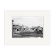 Archival quality photographic print titled Pukekohe 1910 - HI200 featuring a small New Zealand town street with wooden buildings, horse-drawn carriage, utility poles, picket fence, and people near the carriage.