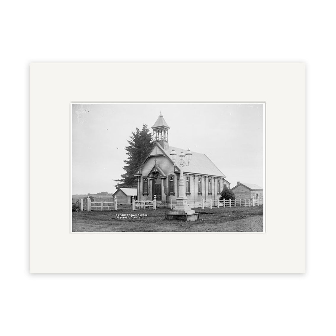 Black and white historical photographic print of St James Presbyterian Church 1915 (HI201), a small wooden church with a bell tower in New Zealand, surrounded by a fence, trees, other buildings, and featuring a sign and bench in front.