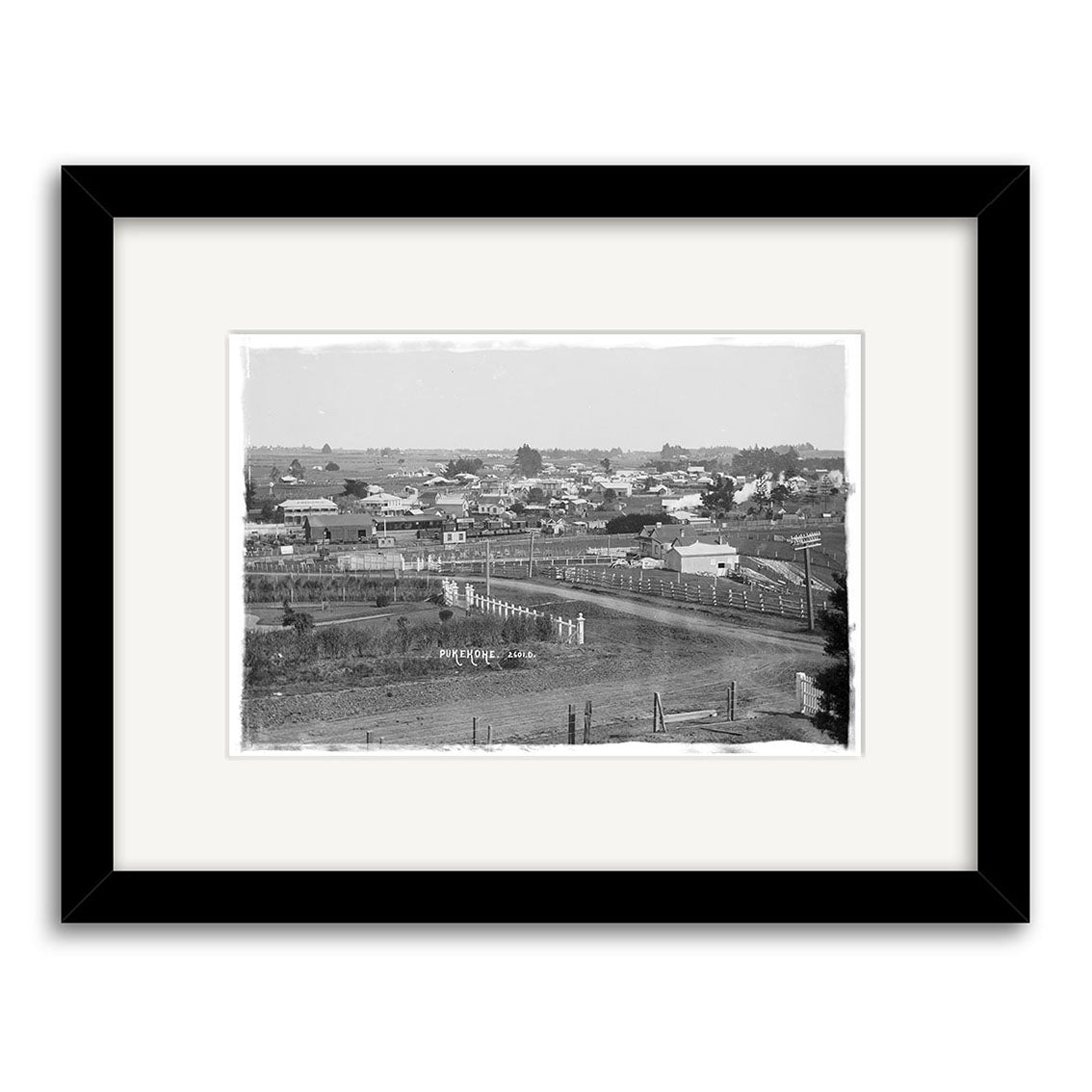 Black and white photograph titled General View Pukekohe 1910 – HI203, depicting a small New Zealand town with scattered houses, dirt roads, and open land. Archival quality print, framed in black with a white mat.