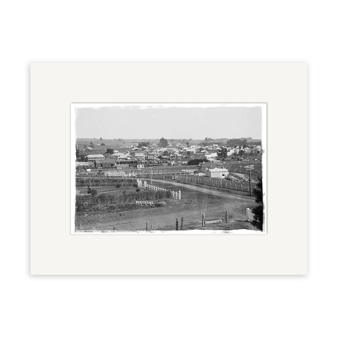 Black and white photograph titled General View Pukekohe 1910 - HI203 shows a small New Zealand town with scattered houses, open fields, and people by a dirt road; the historical print features a wide white mat border.