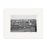 Black and white photograph titled General View Pukekohe 1910 - HI203 shows a small New Zealand town with scattered houses, open fields, and people by a dirt road; the historical print features a wide white mat border.