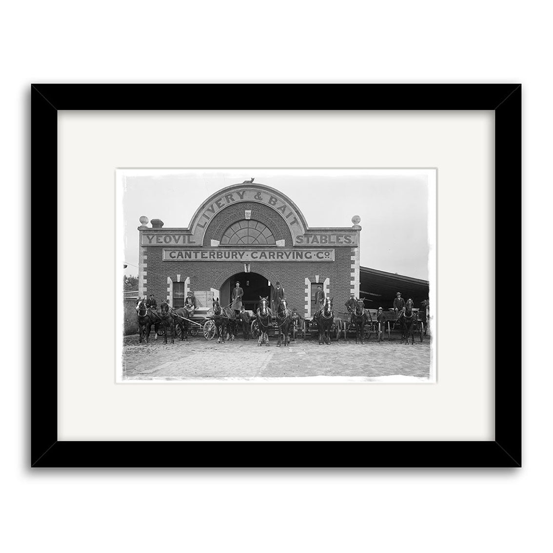 Archival-quality black-and-white photo print of the Canterbury Carrying Company 1910 (HI204), showing a brick building with horse-drawn carriages and people outside; black frame with white mat. Made in New Zealand.