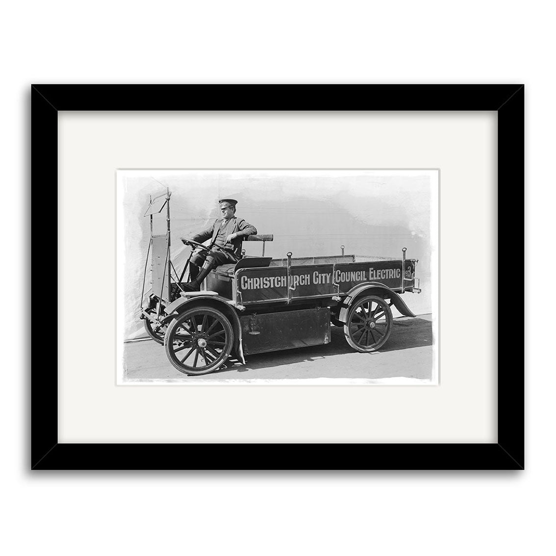 Black-and-white photo of a man in uniform in an early electric vehicle labeled Christchurch City Council Electric, framed with a white mat. Historical Photographic Print from New Zealand: City Council Electric 1920s – HI211.
