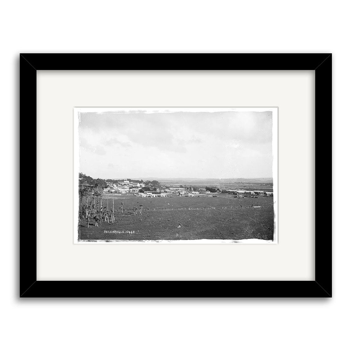The framed black-and-white archival photo Helensville 1914 No.2 - HI215 shows a distant view of a small New Zealand town with sparse buildings on flat open fields beneath a cloudy sky.