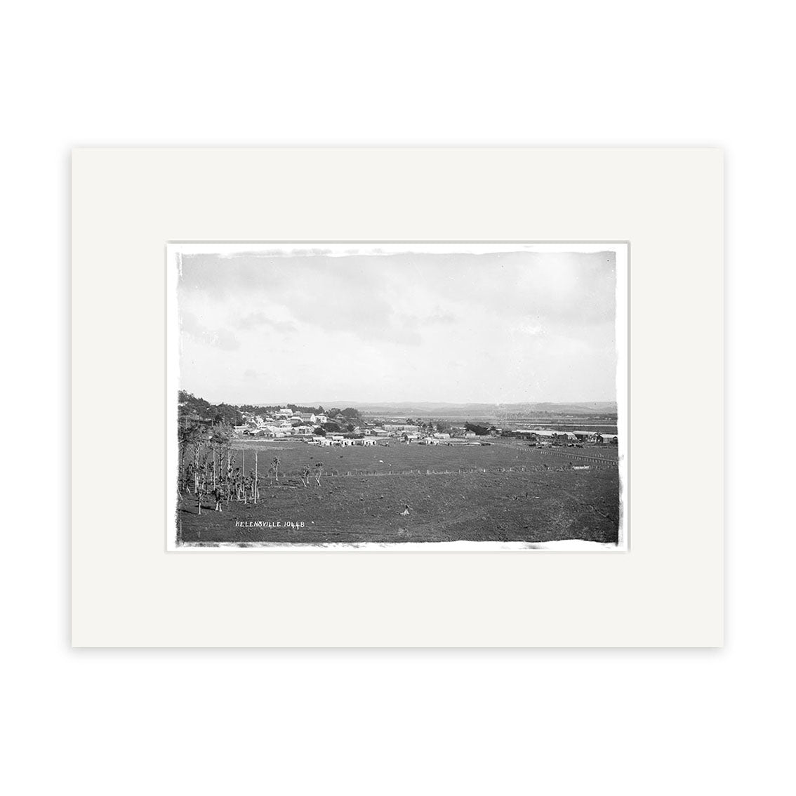 Helensville 1914 No.2 - HI215 is a black and white photographic print of a rural New Zealand landscape with a small town, open fields, and a partly cloudy sky, framed with a wide white mat.