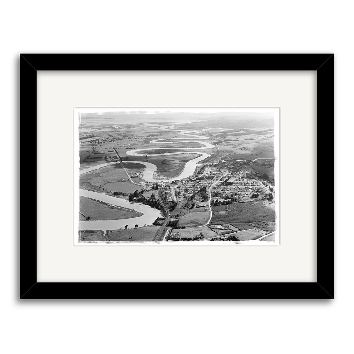 Helensville 1951 - HI216: A black and white aerial photograph of a small town with farmland and a winding river, printed on archival matte paper and presented in a black frame with white mat.
