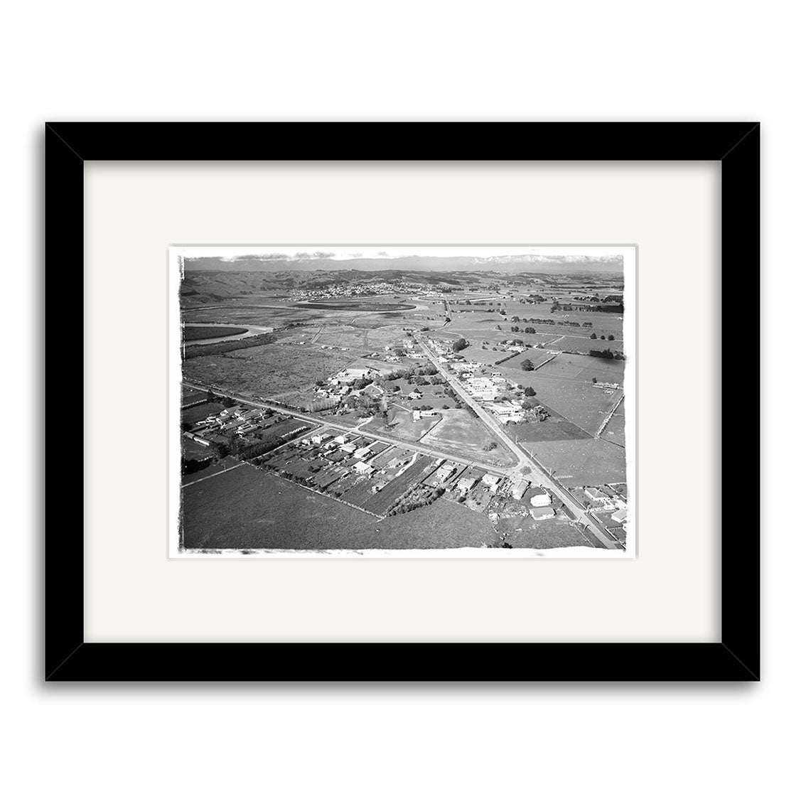 The Helensville 1968 - HI217 is a black and white aerial photographic print on matte paper, presented in a black frame with a white mat. It shows houses, roads, fields, and hills, with archival quality for lasting beauty.