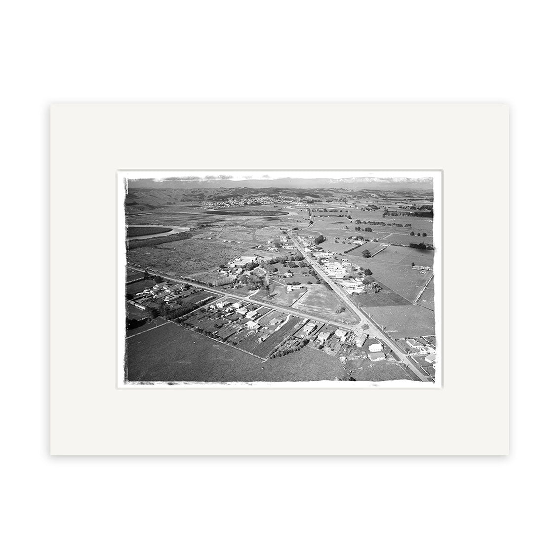 The black-and-white aerial photograph Helensville 1968 - HI217 shows a small rural town with houses, roads, and fields bordered by hills. This archival quality photographic print comes matted with a wide white border.