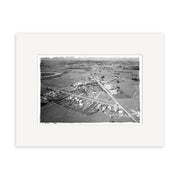 The black-and-white aerial photograph Helensville 1968 - HI217 shows a small rural town with houses, roads, and fields bordered by hills. This archival quality photographic print comes matted with a wide white border.